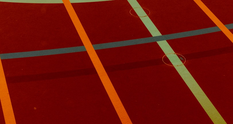 A sports court with intersecting lines in orange and blue, creating geometric patterns on a reddish-brown surface.