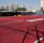 A sports court with a red playing surface and white lines, surrounded by a black mesh fence. In the background, tall buildings are visible, along with some trees and a few yellow chairs positioned near the fence.